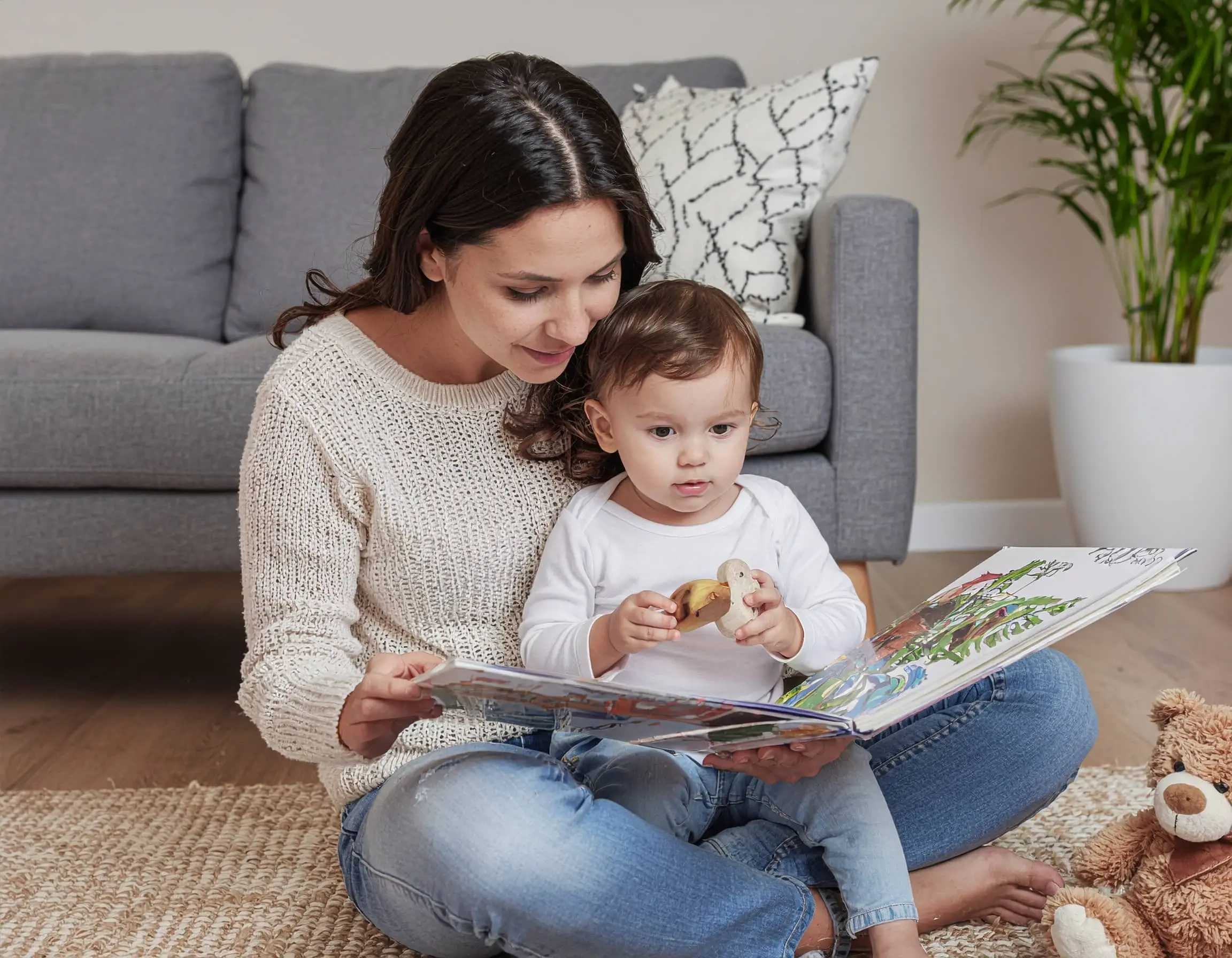 Μαμά διαβάζει βιβλίο σε μικρό παιδί καθισμένες στο πάτωμα του σαλονιού, με λούτρινα παιχνίδια δίπλα τους