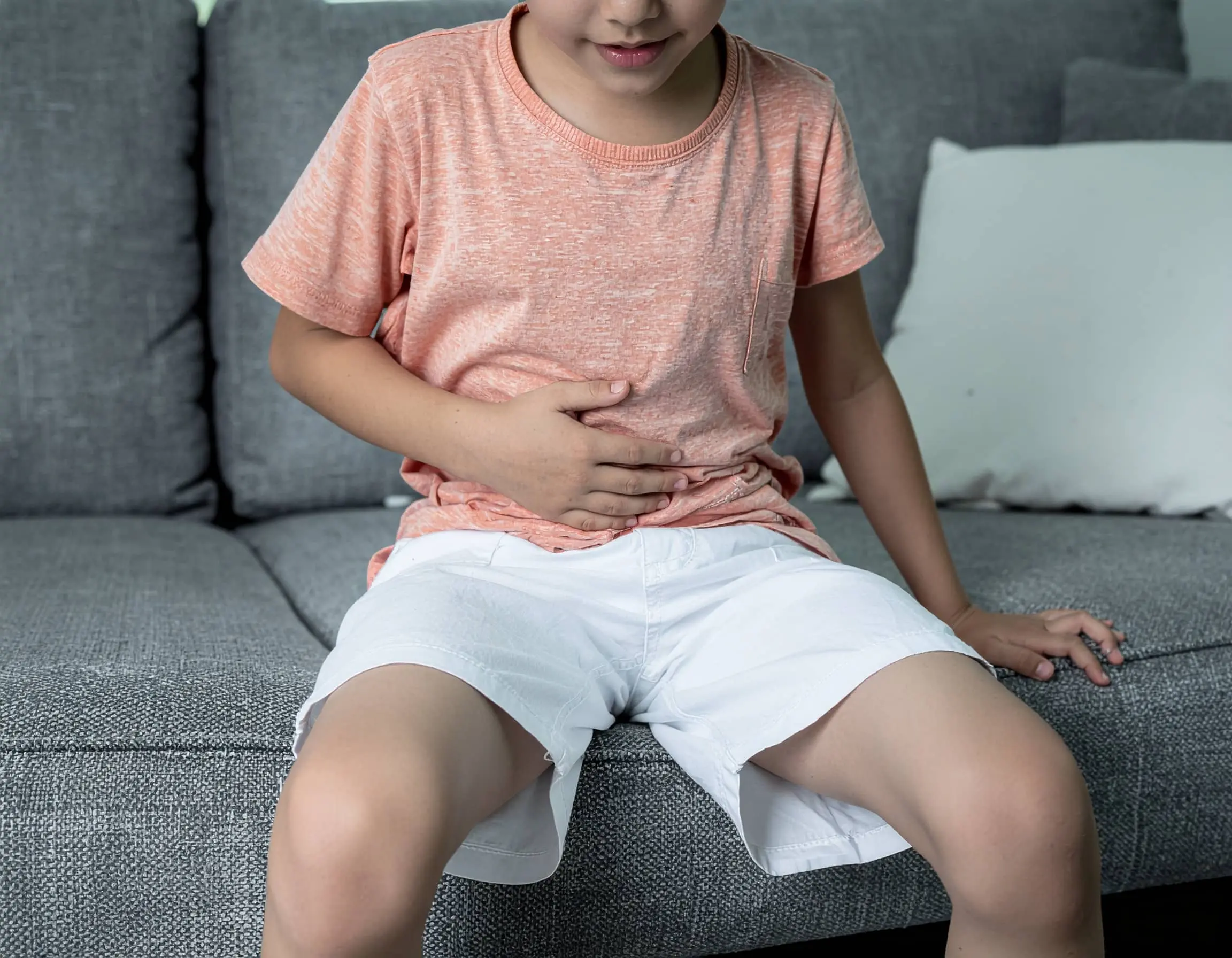 Αγόρι με πορτοκαλί μπλούζα και λευκό σορτς κάθεται σε καναπέ και κρατά την κοιλιά του, δείχνοντας ενόχληση ή πόνο.