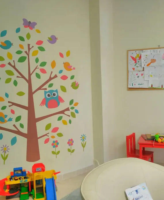 Η παιδίατρος 8 Τοίχος παιδικού χώρου διακοσμημένος με αυτοκόλλητο δέντρο και πουλάκια, παιδικό τραπεζάκι με κόκκινες καρέκλες και παιχνίδια, και πίνακας με ζωγραφιές παιδιών