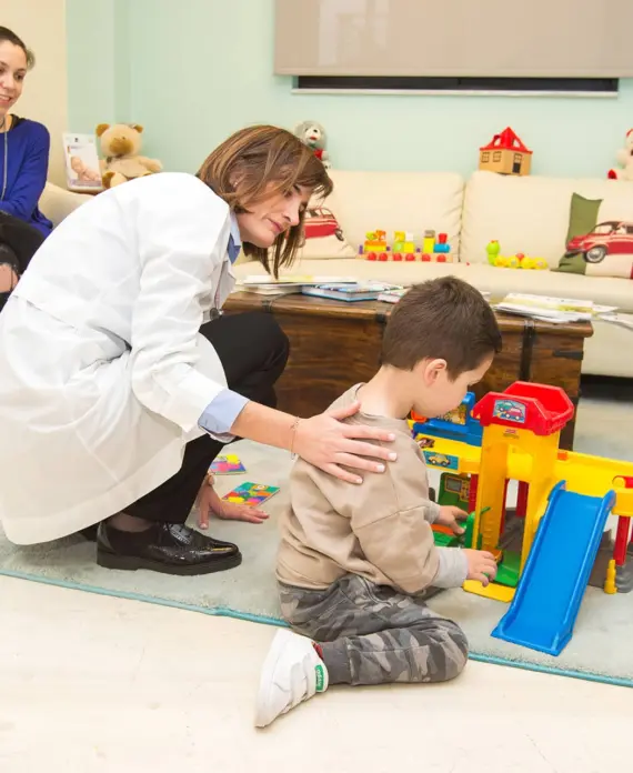 Η παιδίατρος 5 Παιδίατρος γονατιστή δίπλα σε αγόρι που παίζει, με τη μαμά να παρακολουθεί χαμογελώντας στον παιδικό χώρο αναμονής.