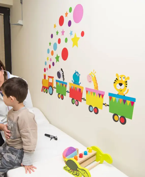 Η παιδίατρος 4 Παιδίατρος εξετάζει αγόρι σε εξεταστήριο διακοσμημένο με πολύχρωμο αυτοκόλλητο τρενάκι και παιχνίδια, δημιουργώντας φιλική ατμόσφαιρα